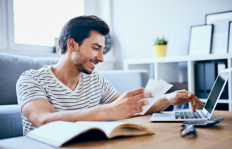 Man smiling while holding receipts and looking at laptop
